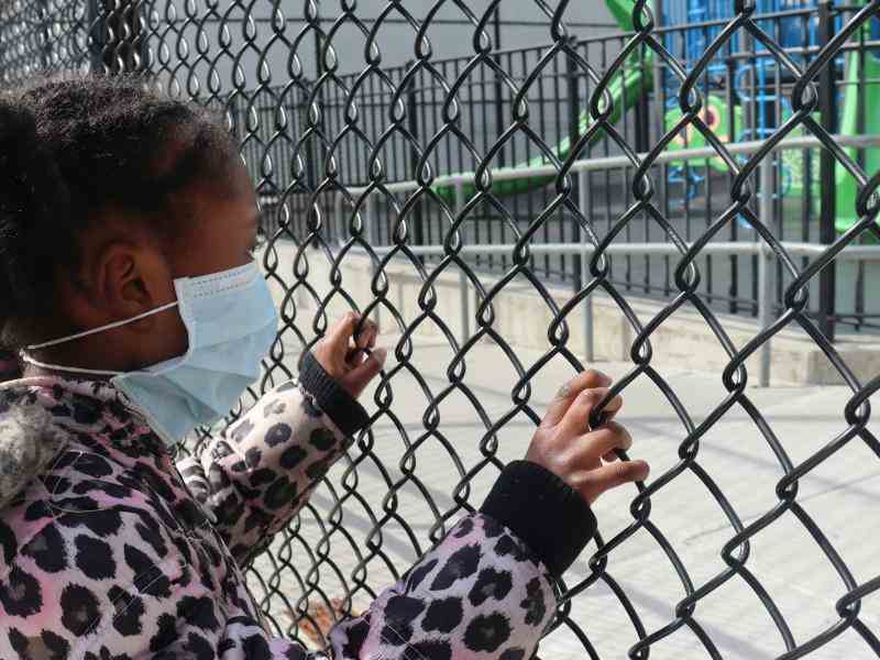 young child with hands on a chain-link fence looking at a playground inside the fence