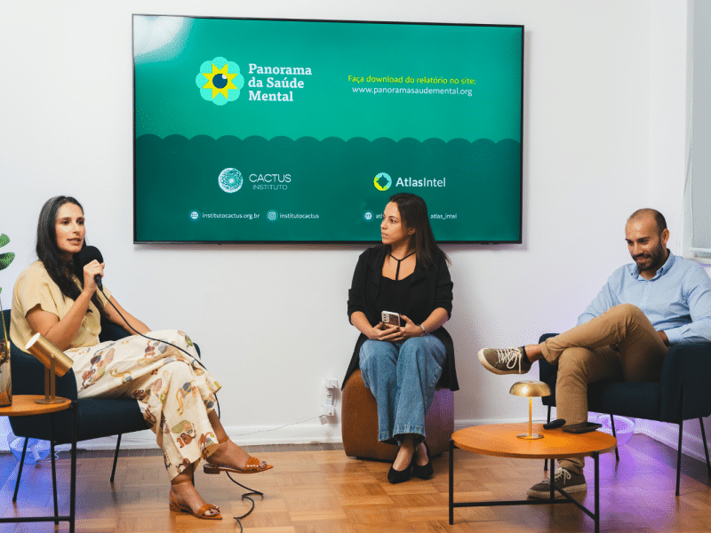 Três pessoas sentadas em poltronas falam em um painel. Atrás delas, há uma televisão que dá destaque ao Panorama da Saúde Mental