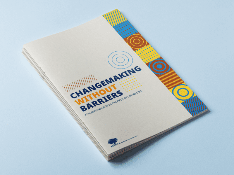 Stack of printed reports titled “Changemaking Without Barriers: Ashoka’s Insights in the Field of Disabilities,” placed on a light blue surface, with a beige cover and a vertical strip of colorful geometric patterns on the right side.