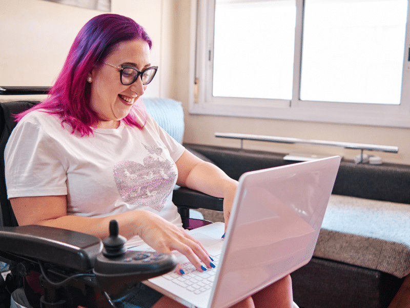 A person with pink hair uses a laptop while seated in a powered wheelchair in a bright living room.