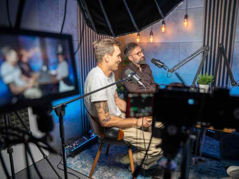 Two man sitting in podcast studio by the table talking to the microphone