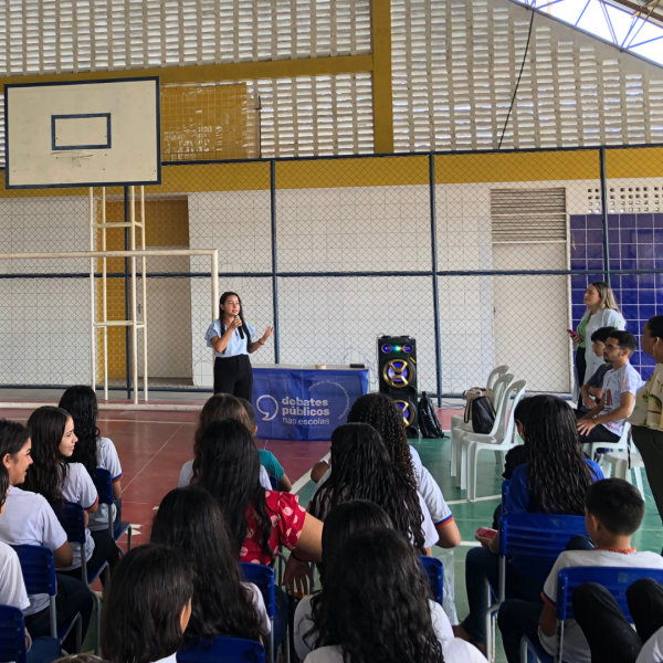 Vê-se uma foto de diversos estudantes olhando uma pessoa falando ao microfone com a bandeira dos Debates Públicos nas Escolas atrás dela