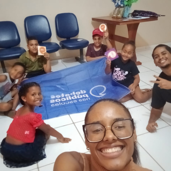 Vê-se uma roda de diversas crianças com Marta (Mulher negra de cabelos pretos e longos usando óculos) e Pedro Pedro (Homem negro de cabelos pretos presos e com as laterais mais curtas com barba no queixo) ao redor da bandeira dos Debates Públicos nas Escolas