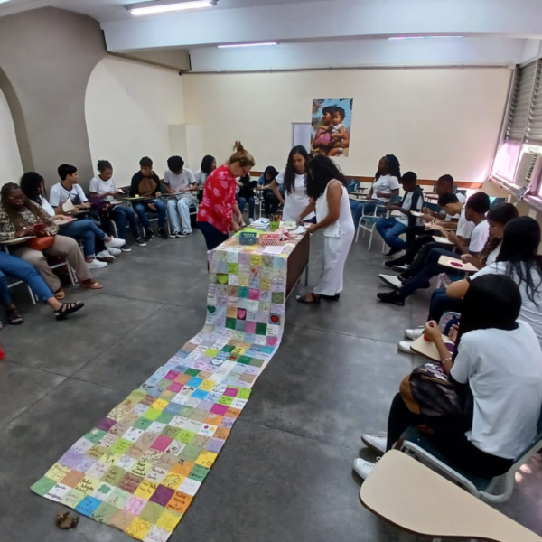 Vê-se uma foto de diversas pessoas sentadas em meia lua com uma mesa e uma grande colcha de retalhos