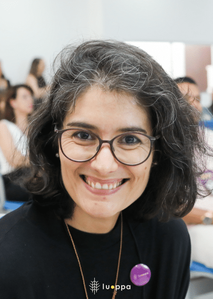 Foto de Mónica, mulher branca com cabelo preto na altura dos ombros e raízes grisalhas. Ela sorri para a foto e usa óculos, uma camiseta preta e um botton roxo. Ao fundo, há pessoas sentadas em cadeiras.