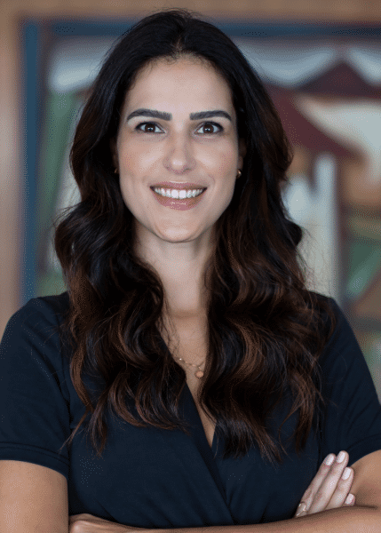 Foto de Carolina, mulher branca de cabelos ondulados, compridos e castanhos. Ela usa uma blusa de manga curta azul marinho e posa para a foto, sorrindo de braços cruzados