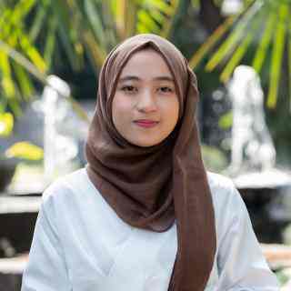 Headshot of AYC Devy smiling directly at the camera wearing a brown hijab and white shirt on a nature background.