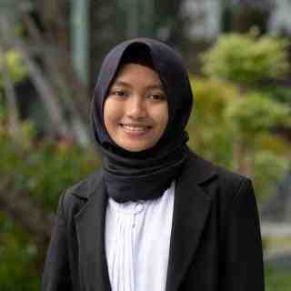 Headshot of AYC Diah smiling directly at the camera wearing a black hijab and a black suit on a nature background.