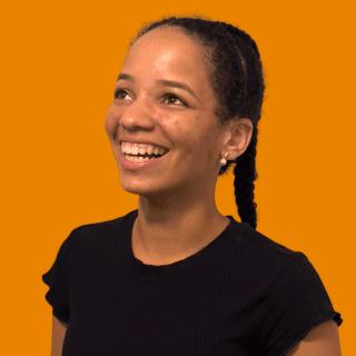 Isabela é uma jovem com cabelos crespos e escuros. Ela usa uma trança e sorri