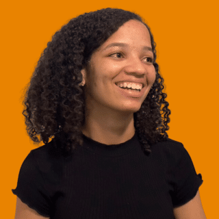 Isadora é uma jovem negra de cabelo médio e cacheado. Ela veste uma blusa preta e sorri