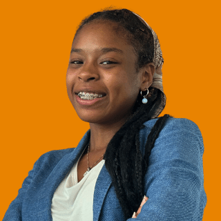 Jahzara é uma jovem negra com tranças longas. Ela veste uma blusa branca com uma camisa azul, está de braços cruzados e sorri para a foto