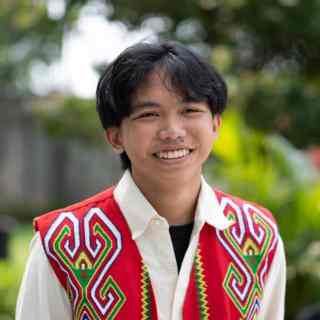 Headshot of AYC Kynan smiling directly at the camera wearing a white shirt and a traditional red vest of Dayak Tribe on a nature background