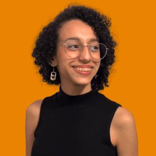 Luísa é uma jovem com cabelos curtos e cacheados. Ela usa óculos e está olhando para o lado sorrindo