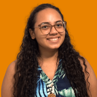 Ruanne é uma jovem com cabelos longos e cacheados. Ela usa óculos, uma blusa estampada e sorri para a foto