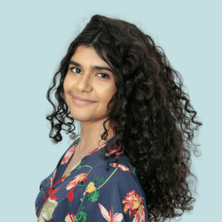 Ana Luiza é uma jovem negra de cabelos longos, escuros e cacheados. Ela veste uma blusa azul com estampa de flores e sorri levemente