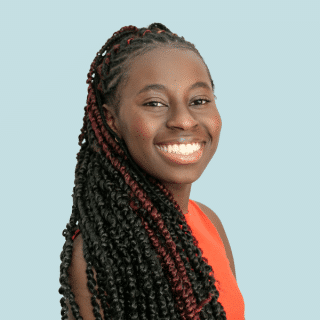 Catarina é uma jovem negra de cabelos escuros e trançados com mexas avermelhadas. Ela sorri para a foto e veste uma regata laranja