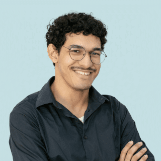 Nicholas é um jovem negro de pele clara com cabelo curto, escuro e cacheado. Ele tem bigode, usa óculos e veste uma camisa social cinza escuro. Nicholas está de braços cruzados e sorri para a foto
