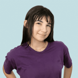 Joana é uma jovem branca de cabelo liso, longo e castanho com franja. Ela usa uma camiseta roxa e sorri levemente para a foto