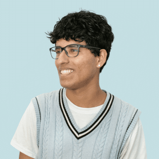 Lucas é um jovem negro com cabelo curto e ondulado. Ele usa óculos, veste uma camiseta branca com um colete de lã azul claro e sorri olhando para o lado
