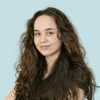 Paula é uma jovem branca de cabelo ondulado, longo e castanho. Ela sorri levemente e olha para a câmera
