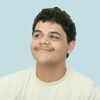 João é um jovem pardo de cabelos curtos, escuros e cacheados. Ele veste uma camisa bege e olha para o lado