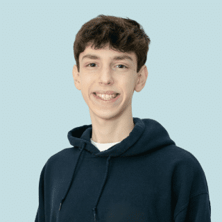 Eli é um jovem branco de cabelos curtos e castanhos. Ele veste um moletom com capuz azul marinho e sorri para a foto