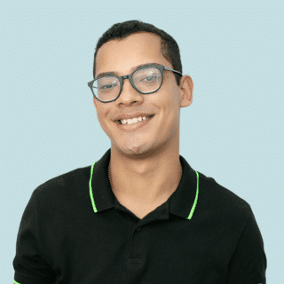 Felipe é um jovem pardo de cabelo curto e escuro. Ele usa óculos, uma camisa polo preta com detalhe verde na gola e sorri para a foto