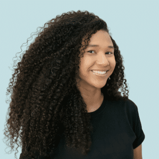 Hagata é uma jovem negra de cabelos cacheados, escuros e volumosos. Ela veste uma camiseta preta do Projeto Boyebi e sorri para a foto