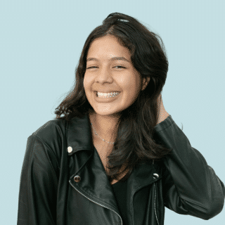 Karollyny é uma jovem branca de cabelo médio, ondulado e castanho. Ela põe a mão no cabelo e sorri para a foto. Karollyny veste uma blusa preta e uma jaqueta de couro preta