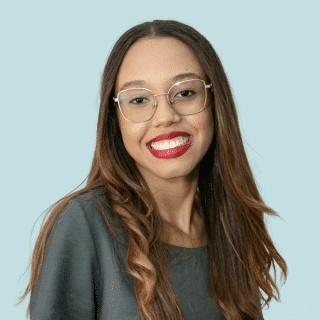 Sabrina é uma jovem negra de cabelo castanho, liso e comprido. Ela usa óculos, um batom vermelho, blusa cinza, e sorri para a foto